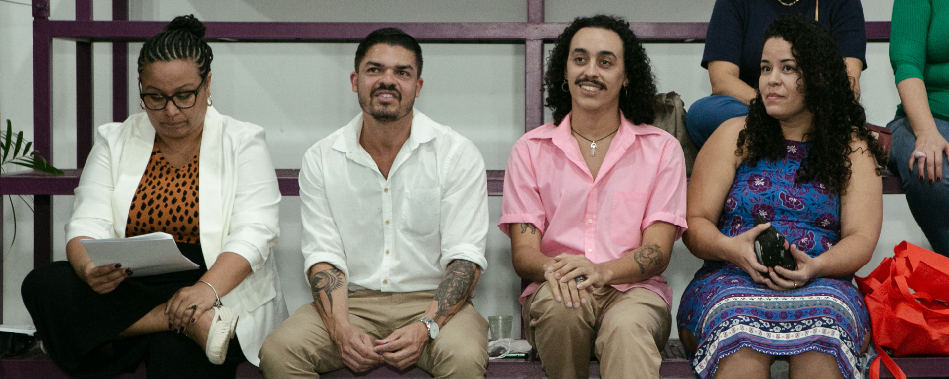 Quatro pessoas estão sentadas - duas mulheres (nas extremidades) e dois homens. Os homens estão sorrindo.