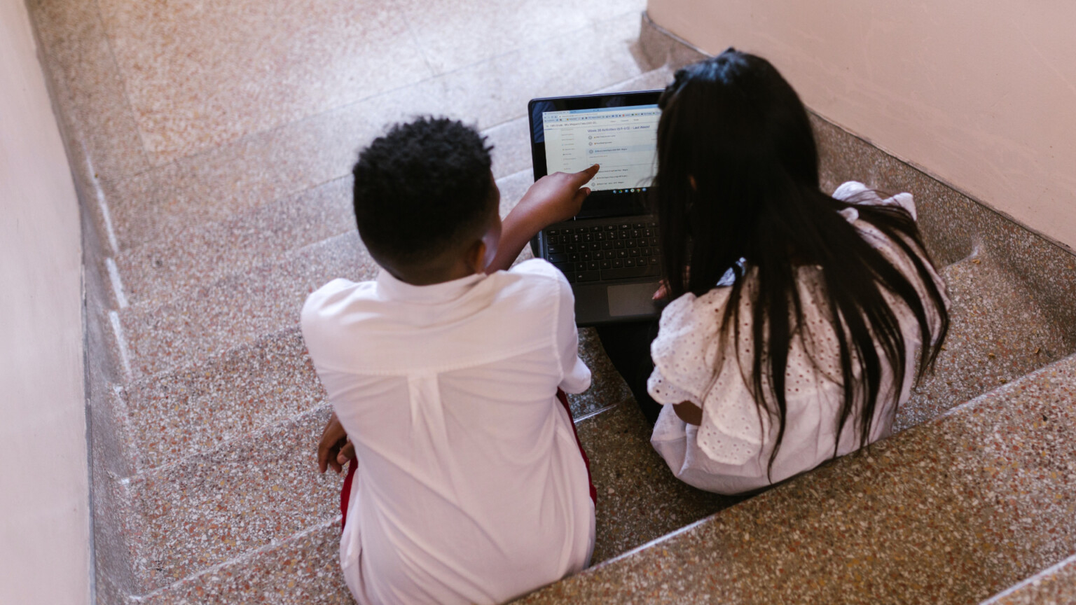 Duas crianças, um menino e uma menina, estão sentados em um degrau de uma escada, de costas para quem os fotografou, enquanto mexem em um pequeno laptop.