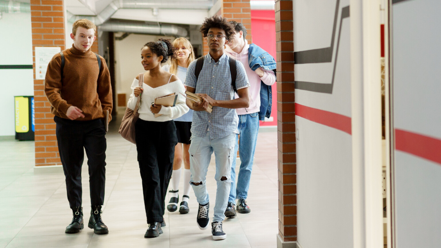 Alguns jovens caminham por um corredor de um ambiente que pode ser uma escola ou universidade.