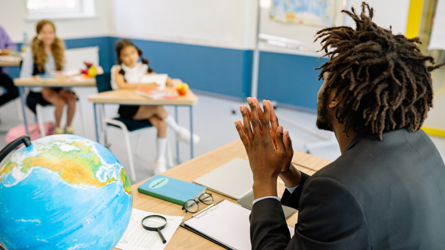 Um professor está sentado à sua mesa, diante da turma de estudantes com idade em torno de 8, 9 anos.