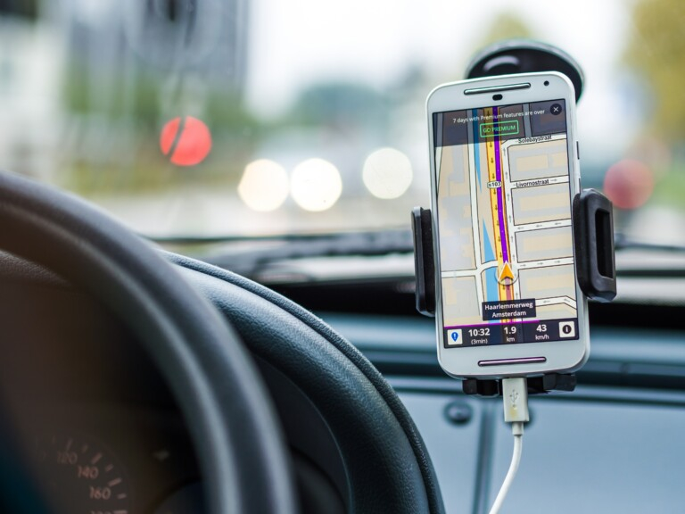 Painel de um carro em que vemos uma parte do volante e do parabrisa. Colado no parabrisa, um suporte de celular com o aparelho mostrando um mapa.