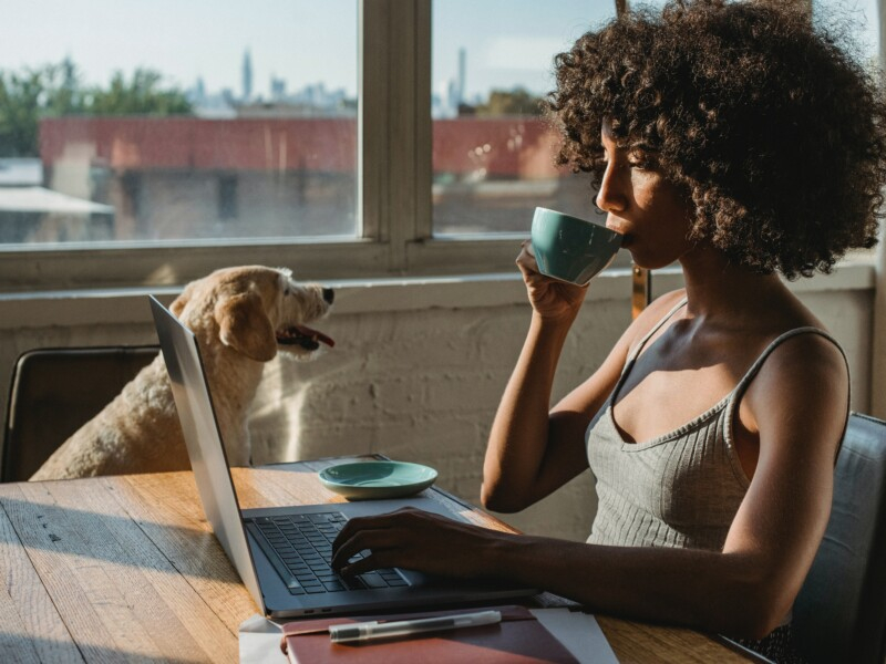 Uma mulher toma um café diante de um laptop, com um cachorro ao seu lado.