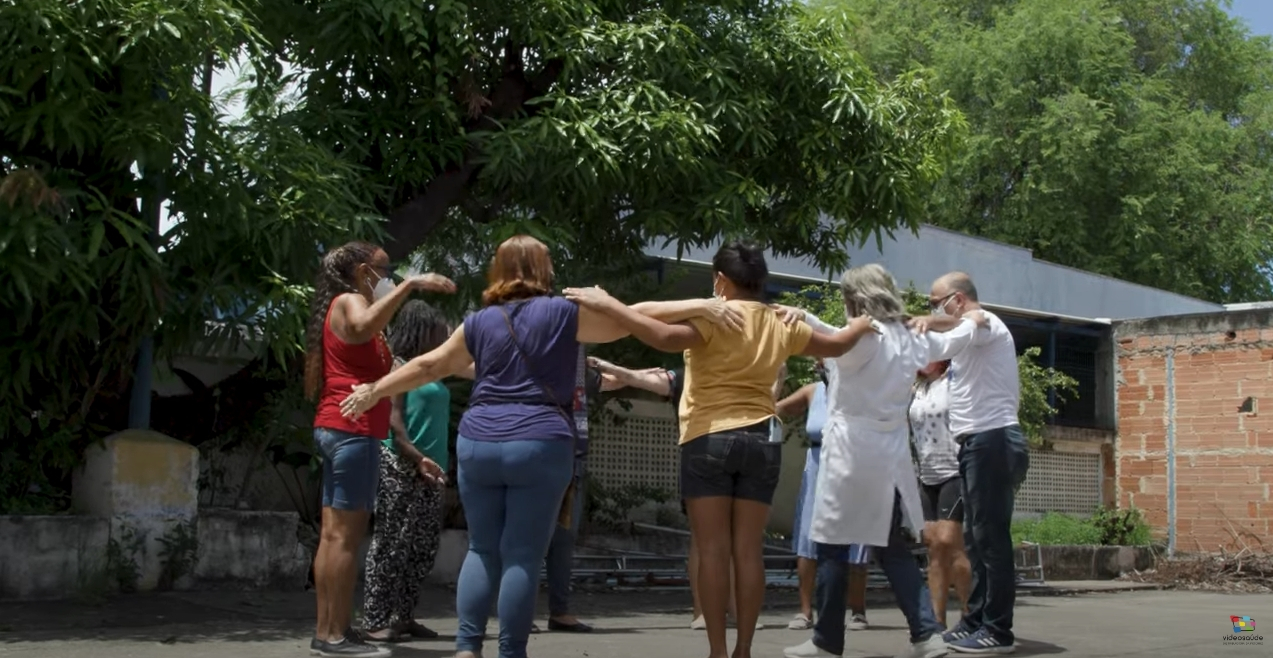 Várias pessoas estão de pé, em círculo, abraçadas. Ao fundo, uma casa e árvores.