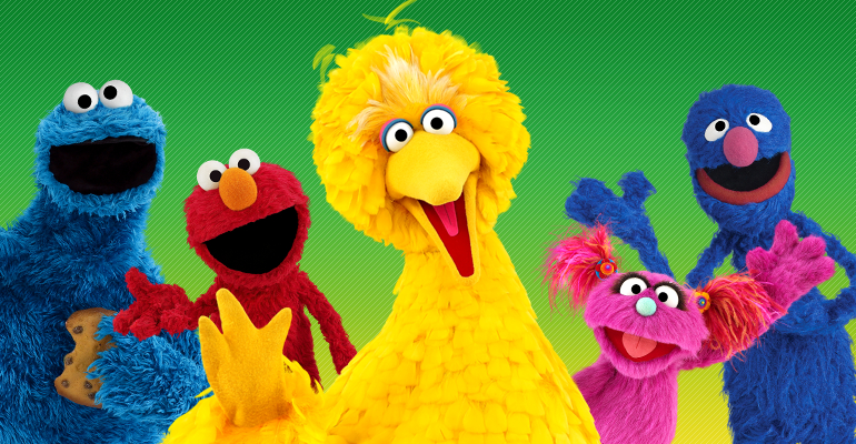 Cinco personagens coloridos que remetem a animais (marionetes ou pessoas fantasiadas) sorriem e acenam.