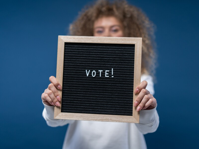 A imagem mostra uma mulher segurando uma placa com "Vote!"