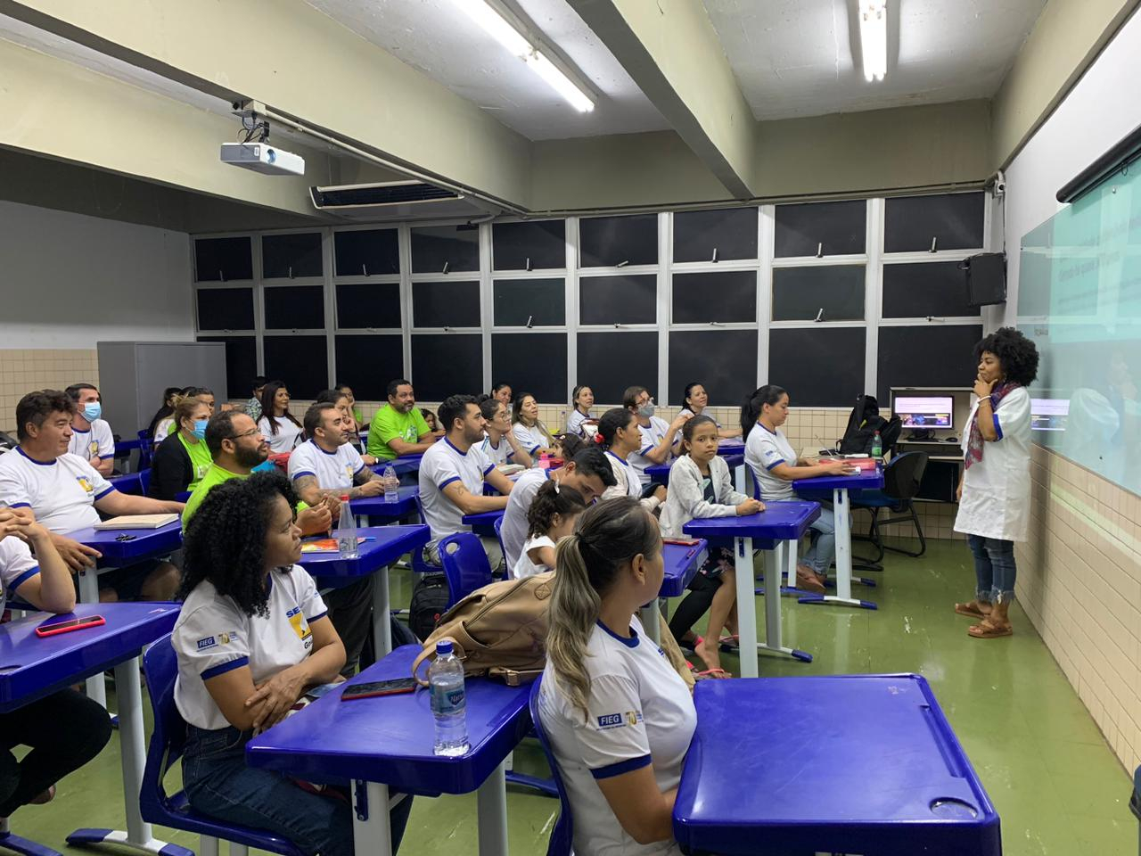 Em uma sala de aula, estudantes estão sentados em suas carteiras, diante da professora.