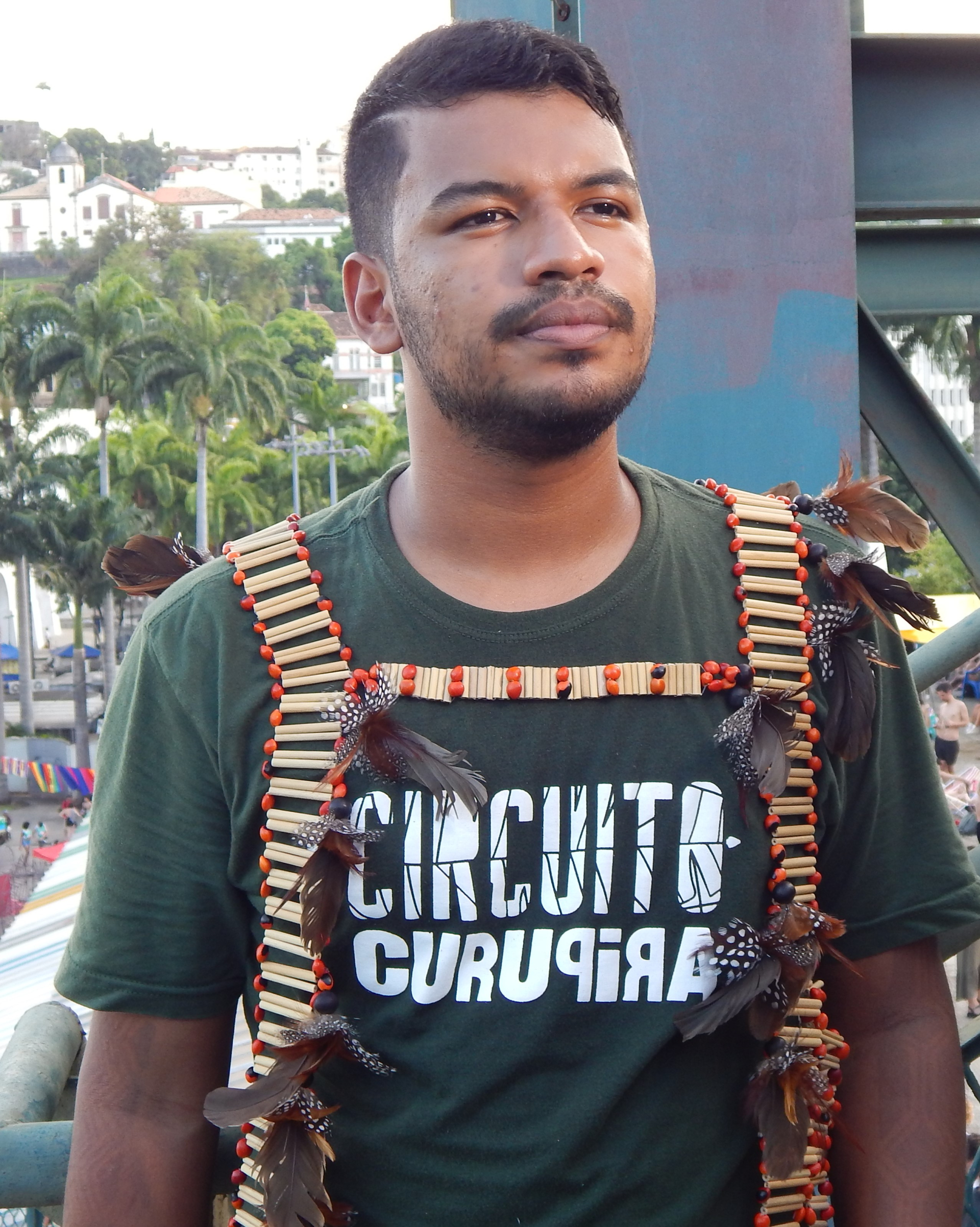 Um homem usa camisa verde-escuro e uma mochila. Ao fundo, árvores e uma estrutura metálica.