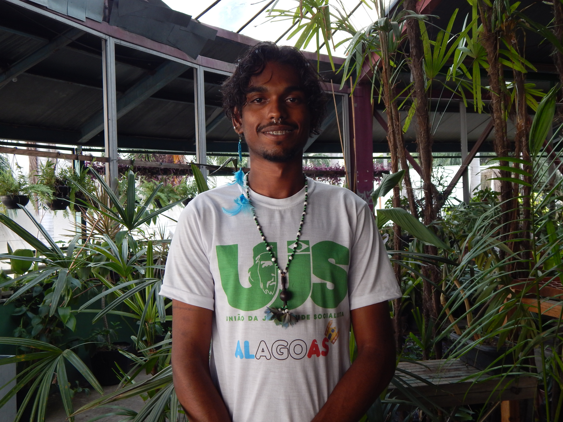 Jovem está de pé, sorrindo levemente, usando uma camisa branca. Ao fundo, plantas.
