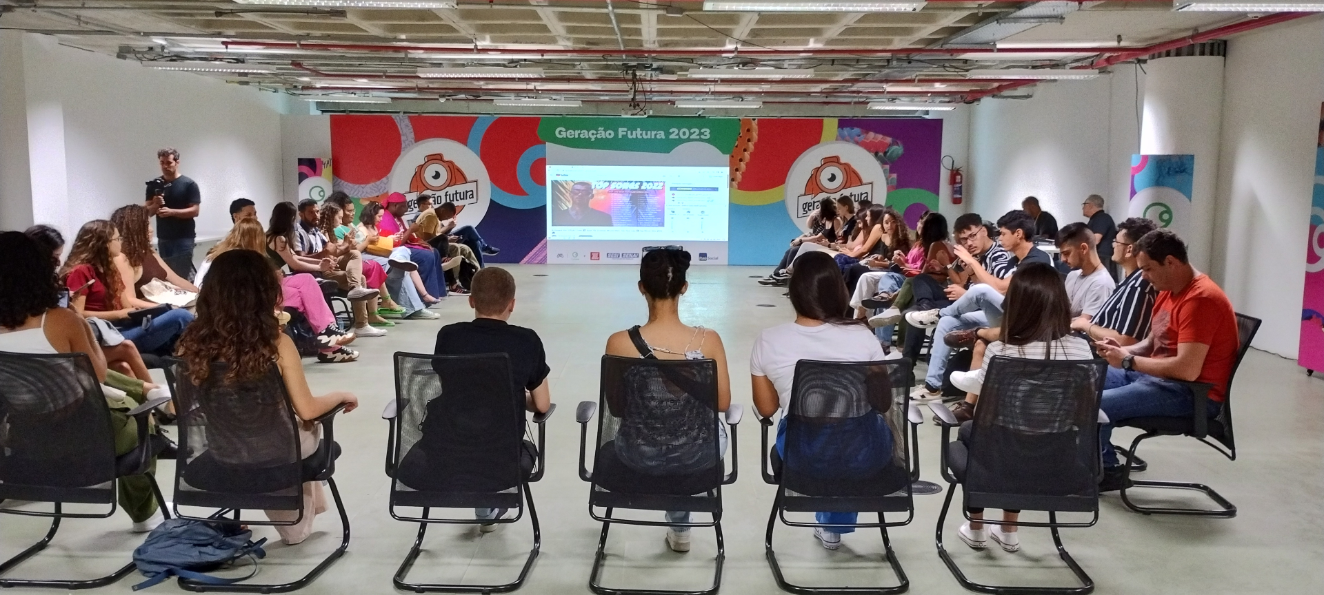 Jovens aparecem sentados, diante de uma tela projetada, de costas pra quem os fotografou.