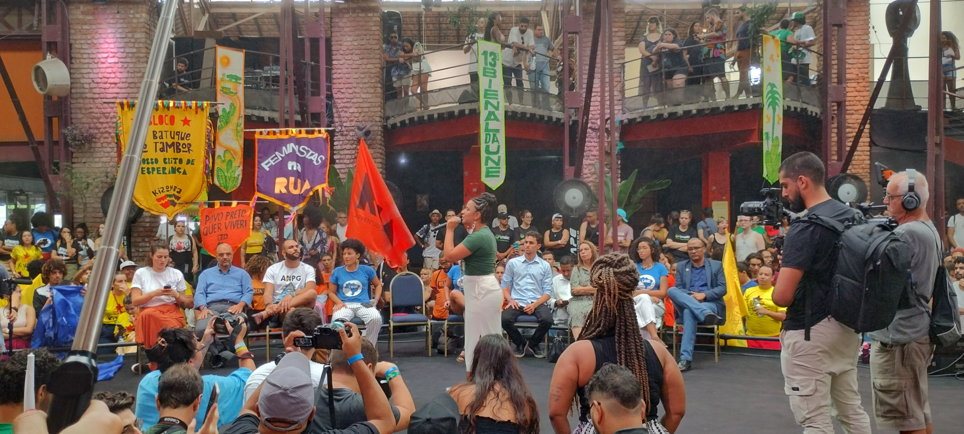 Público assiste ao debate de algumas pessoas que estão sentadas em cadeiras sobre um palco. Há bandeiras ao redor do palco.