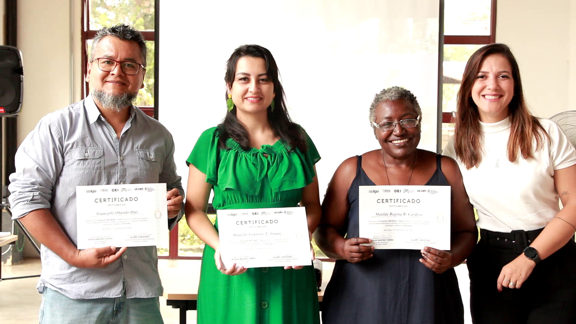 Quatro pessoas estão de pé, lado a lado, sorrindo. Três delas seguram certificados por terem concluído uma etapa do programa Recriar.