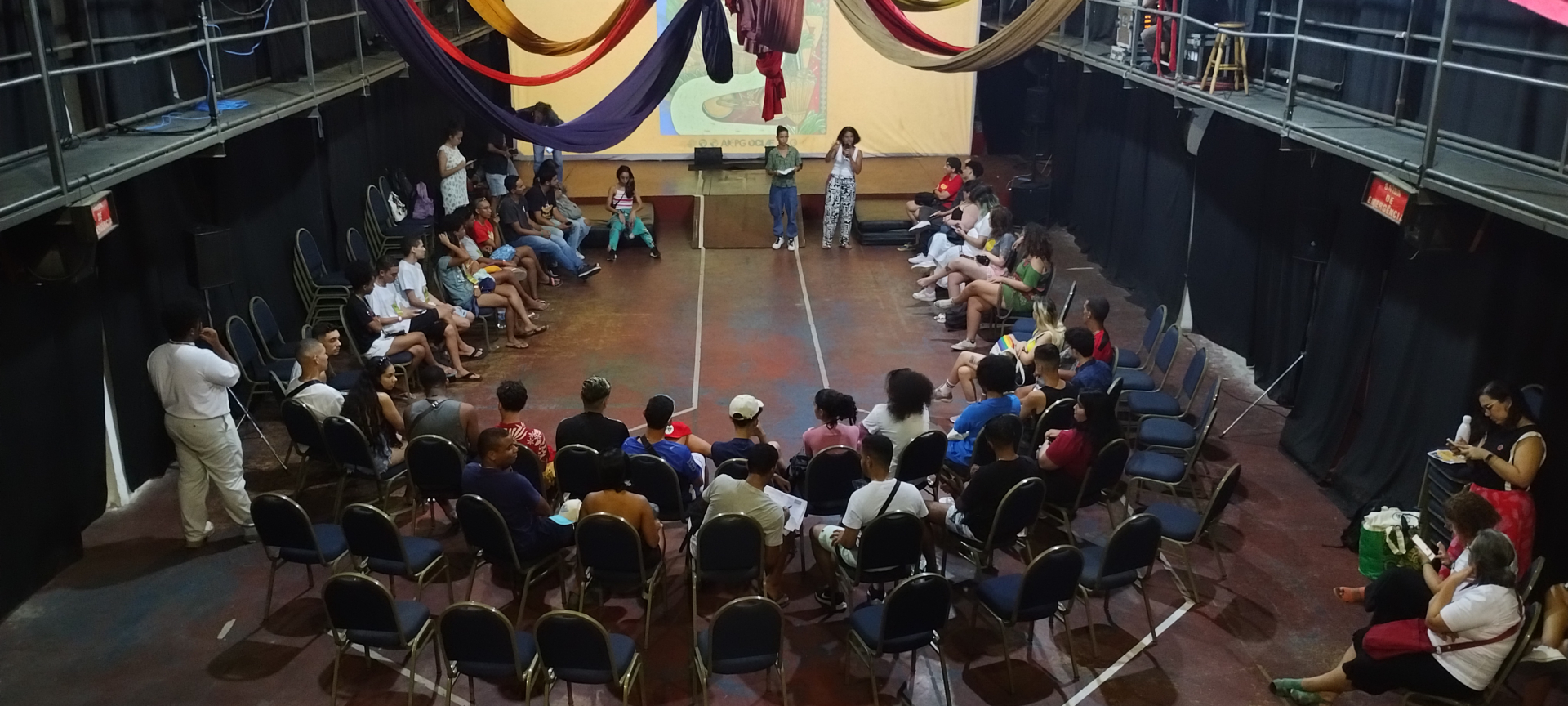 Diversas pessoas sentadas em "u", em um galpão.