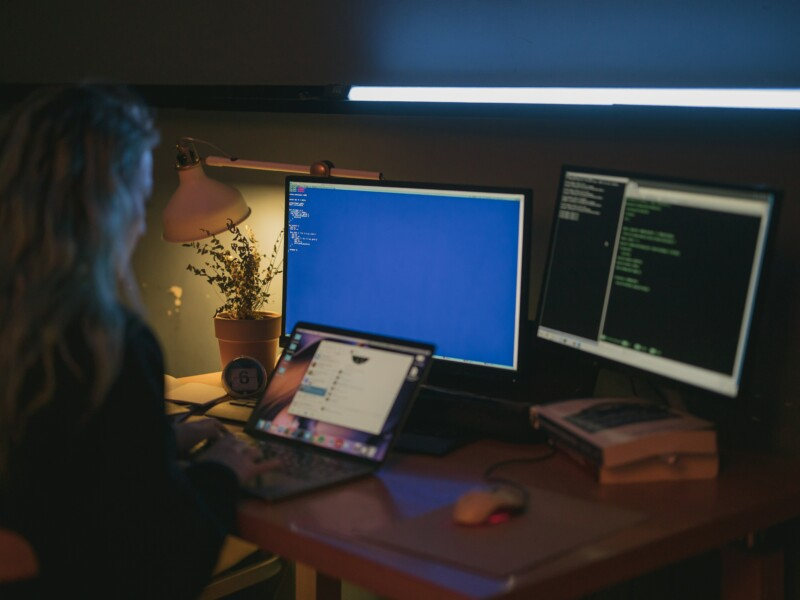 Uma mulher está em uma sala um pouco escura, diante de três telas de computador.