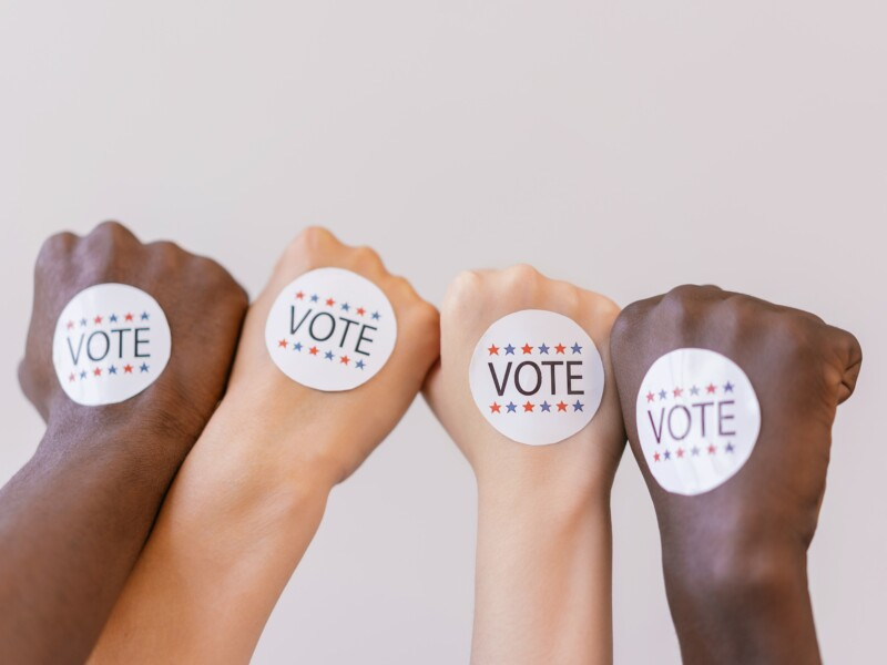 Quatro mãos estão erguidas, de punho fechado, com um adesivo escrito "Vote" em cada uma delas.