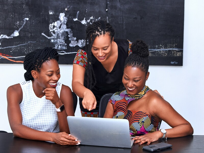 Três mulheres sorriem enquanto olham para a tela do laptop que está diante delas. Duas delas estão sentadas, enquanto uma está de pé.