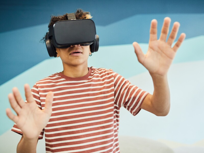 Um jovem está usando óculos de realidade virtual. Ele está com as mãos erguidas, como se estivesse querendo tocar algo.