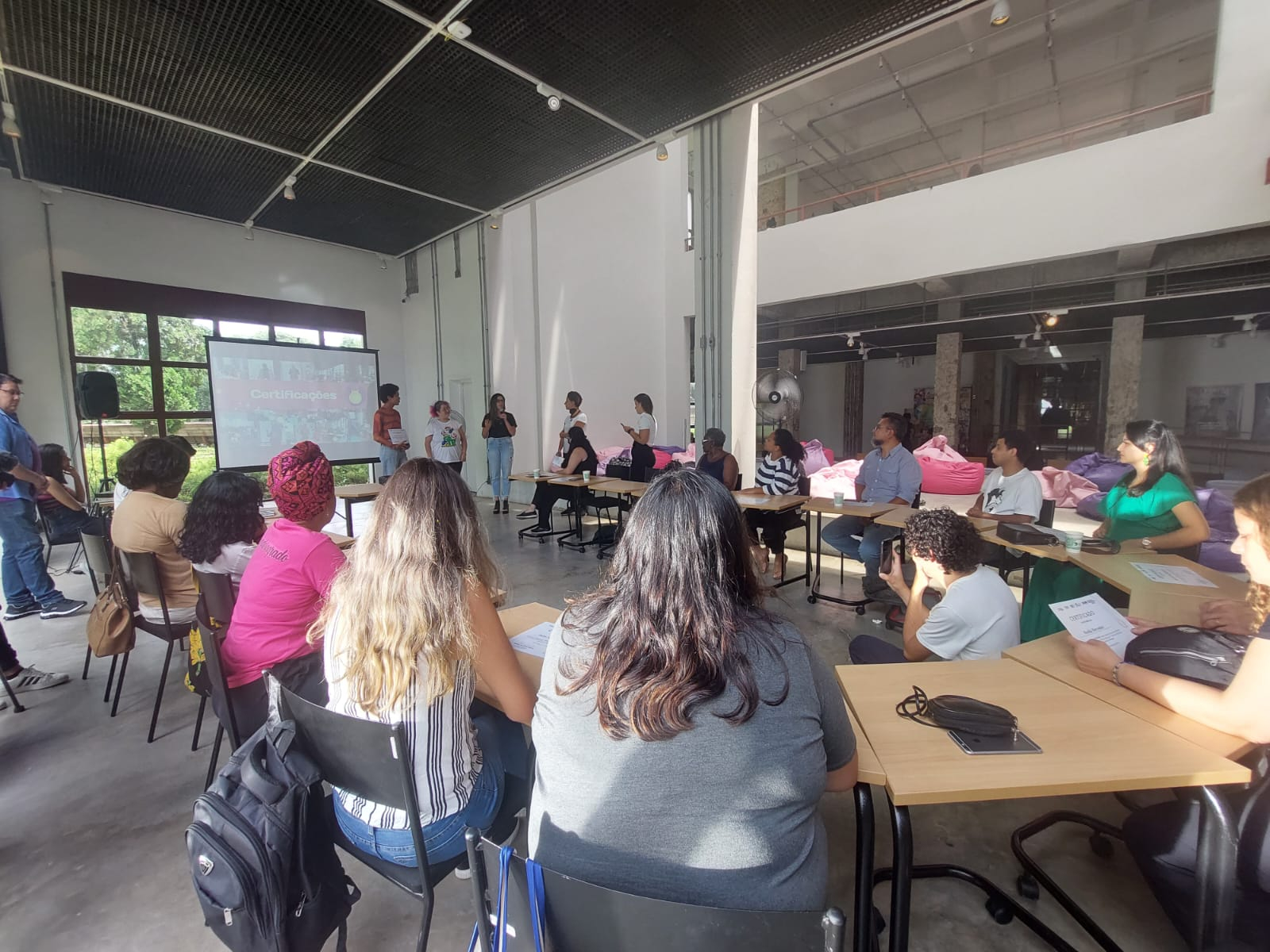 Uma sala em que pessoas estão sentadas em "u", ao redor de uma pessoa em pé.