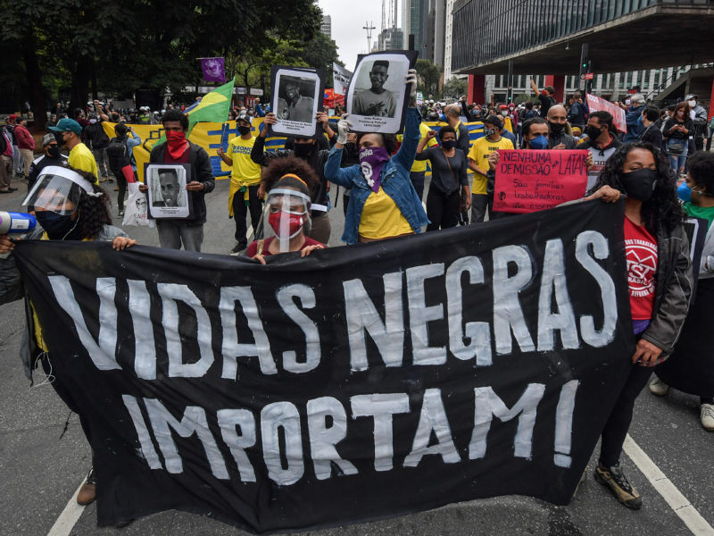 Imagem de um protesto brasileiro. Dezenas de pessoas estão na rua segurando fotos, faixas e cartazes e o que está mais à frente da câmera que fez a foto diz "Vidas Negras Importam"