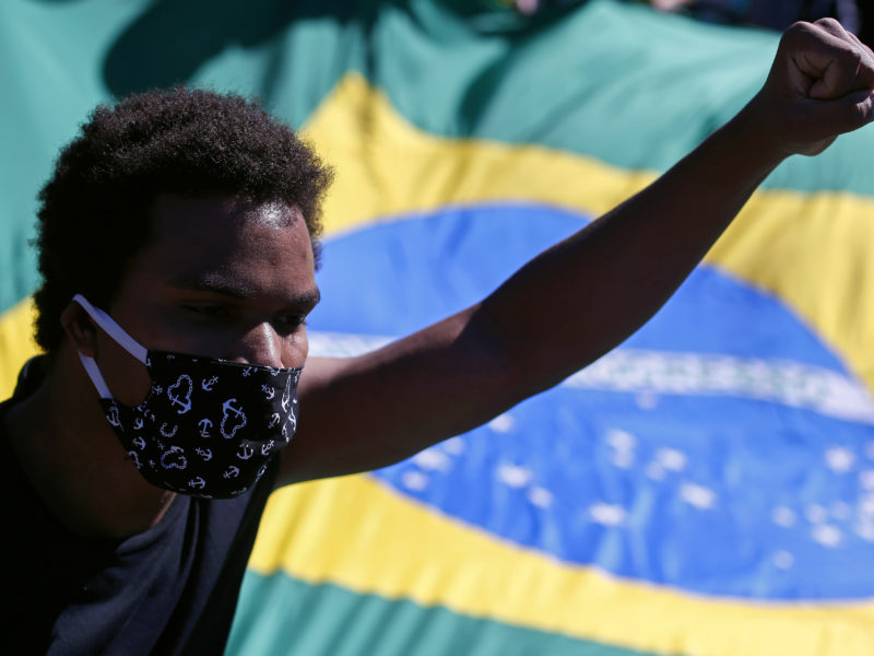 Um jovem negro está de braço esquerdo para cima e punho fechado. Ele está usando máscara de prevenção à Covid-19 e ao fundo há uma bandeira do Brasil