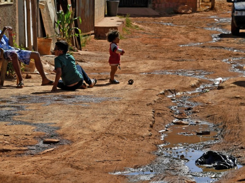 Imagem de uma estrada de barro, enlameada e com duas crianças no centro. Uma é um menino e está sentado no barro e outra é uma menina de uns 3 anos, negra e em pé