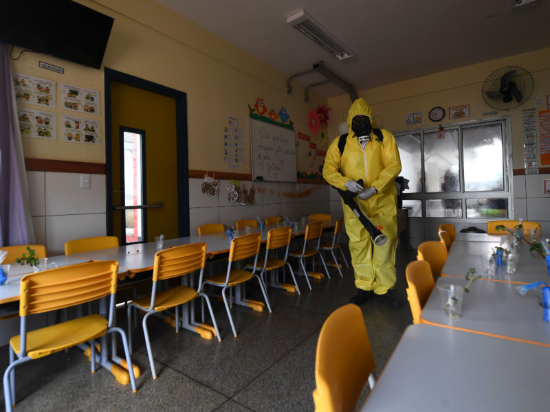 Imagem de uma sala de aula vazia sendo imunizada contra a Covid-19 por uma pessoa trajando uma roupa toda amarela, cobrindo 100% do corpo, incluindo usando máscara no rosto inteiro
