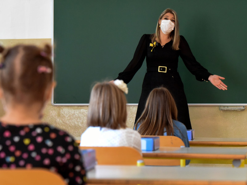 Uma professora de frente para seus alunos sentados em sala de aula e de frente para quem bateu a foto. Ela está gesticulando os braços e parece falar com as crianças, mas está de máscara de prevenção à covid no rosto
