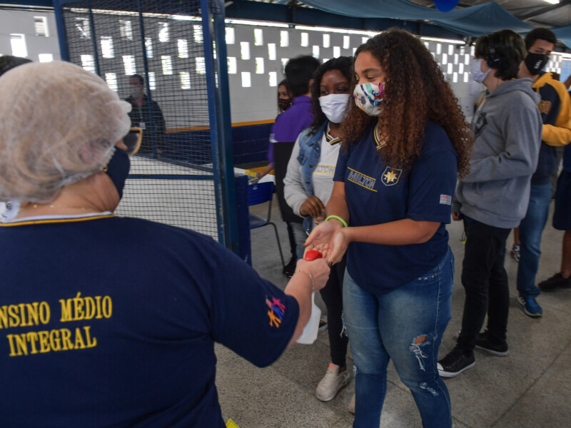 Uma fila de estudantes de máscaras de prevenção à covid-19 está formada em frente à entrada de uma escola. A primeira menina da fila está tendo a temperatura verificada por uma adulta