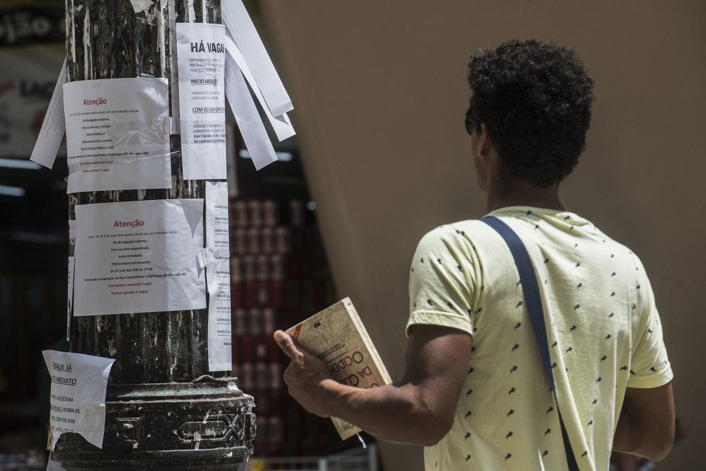 Um homem negro de costas para quem bateu a foto está olhando cartazes de vagas de emprego colados em um poste de rua