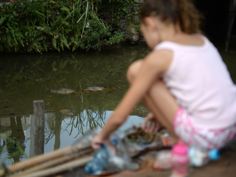 Imagem de uma menina branca agachada e de costas para quem bateu a foto. A menina está de frente para um rio, onde nada um pequeno jacaré