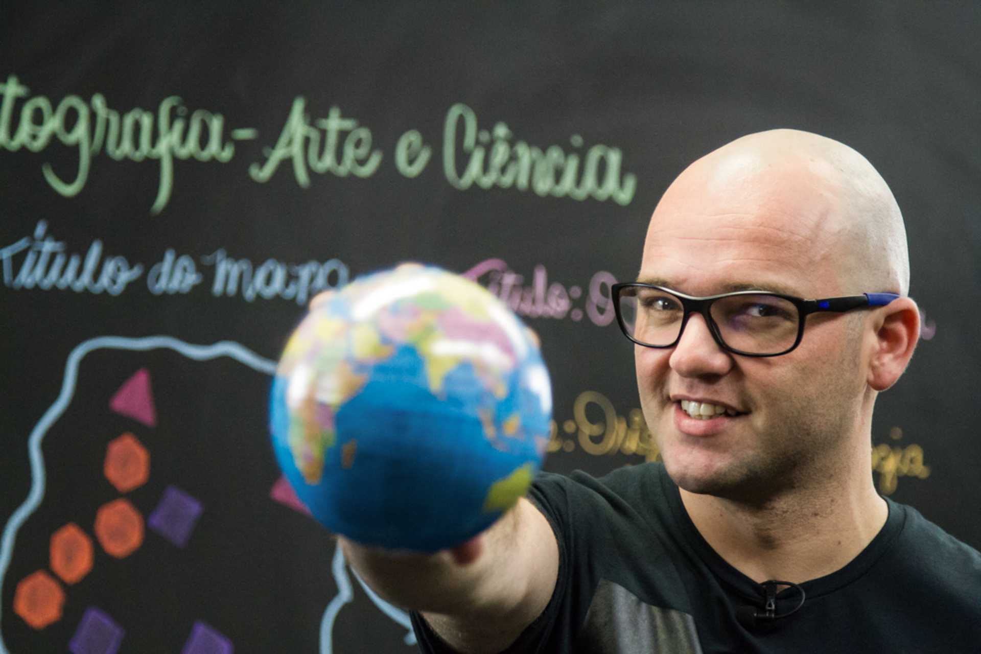 Imagem de um homem branco, careca e de óculos de grau com armação preta. Ele está de camisa cinza escura, segurando um pequeno globo mundial com a mão direita