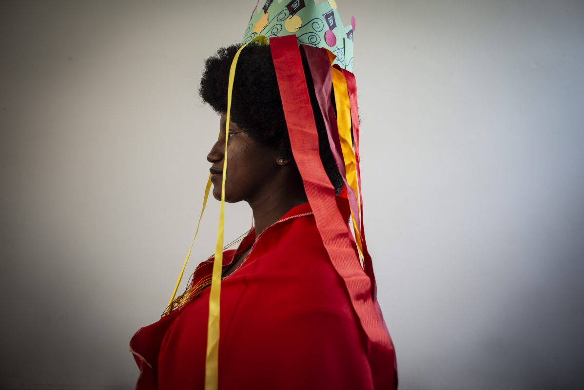 Uma mulher está de lado para quem a fotografou, vestindo uma roupa vermelha, com uma espécie de coroa sobre a cabeça, de onde saem fitas coloridas.