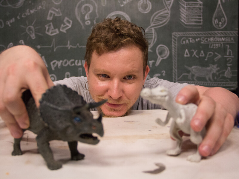 Um homem branco está agachado e brincando com dois dinossauros em uma bancada. Em sua mão direita segura um tricerátops e na mão esquerda um tiranossauro