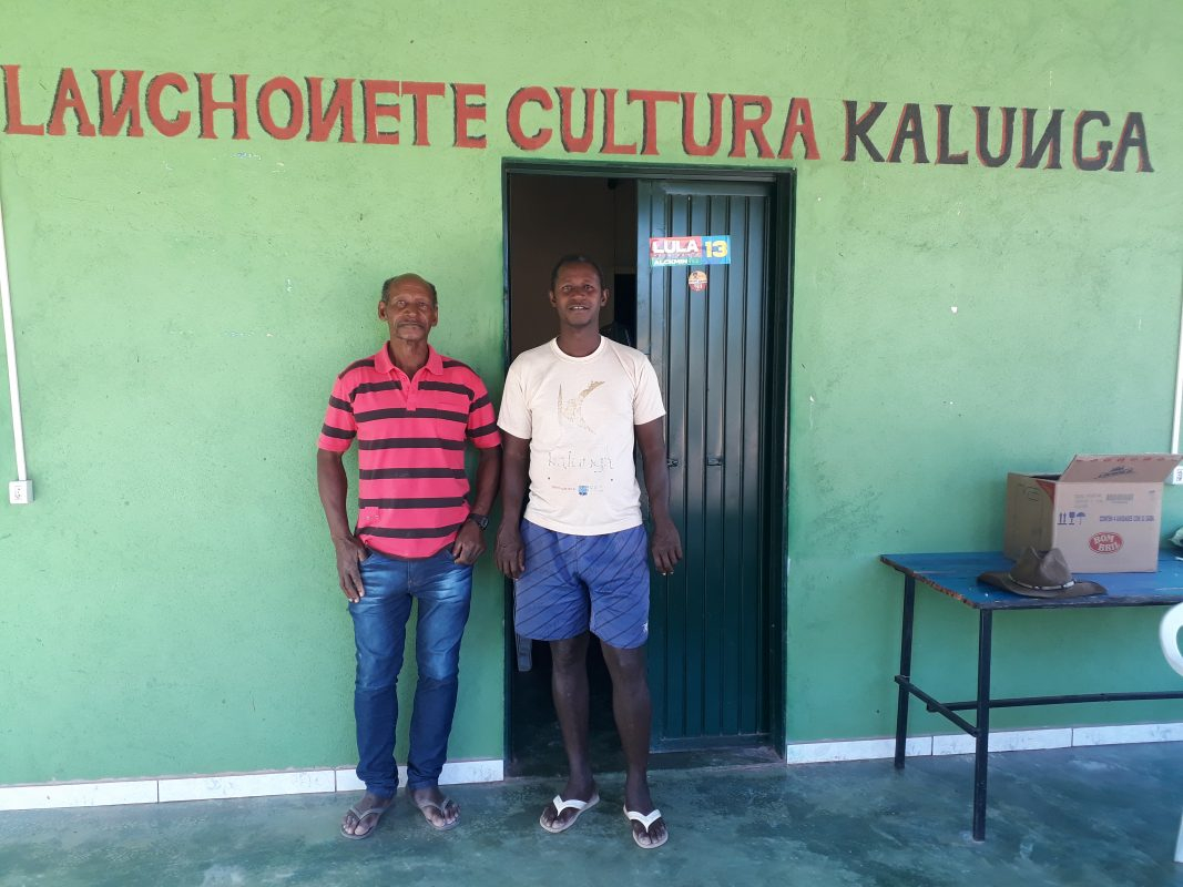 Dois homens estão de pé, de frente para quem os fotografou. Atrás deles, uma parede verde com o nome do estabelecimento escrito na parte superior: "Lanchonete Cultura Kalunga".