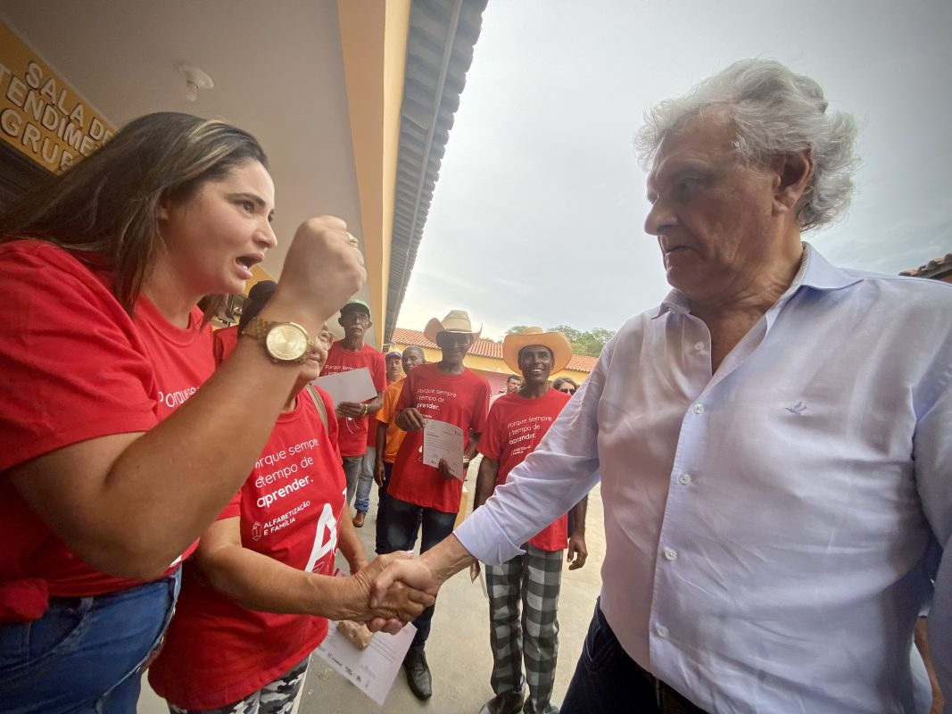 À direita, um homem de cabelo e camisa brancos aperta a mão de um homem de camisa vermelha. No plano anterior ao do homem de vermelho, uma mulher, também de camisa vermelha, gesticula em apoio ao homem de branco.