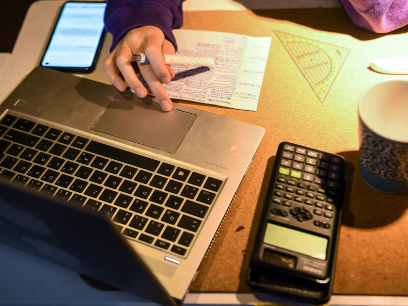 A imagem mostra a mão direita de uma mulher branca mexendo em um notebook aberto sobre uma mesa, que contém outros acessórios, como calculadora, celular e uma folha, por exemplo