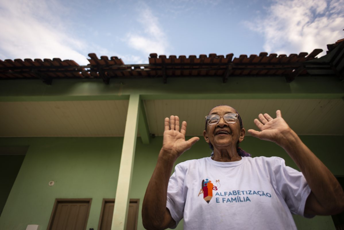 Uma senhora, que usa óculos, sorri com as mãos abertas ao lado de seu rosto. Ao fundo, uma casa e o céu azul.