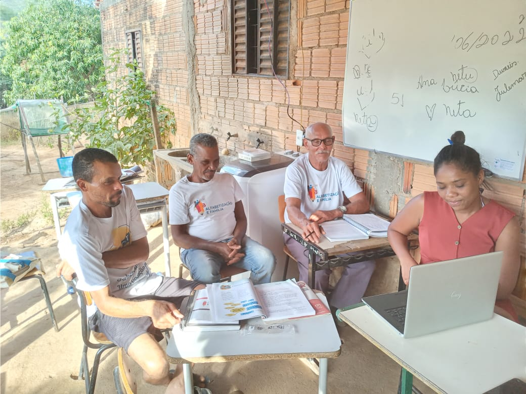 Quatro pessoas estão sentadas na área externa de uma casa, diante de livros e um laptop.