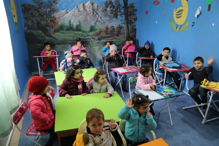 Imagem de uma sala de aula do Jardim de Infância, pois várias crianças pequenas estão sentadas em cadeiras e mesinhas coloridas