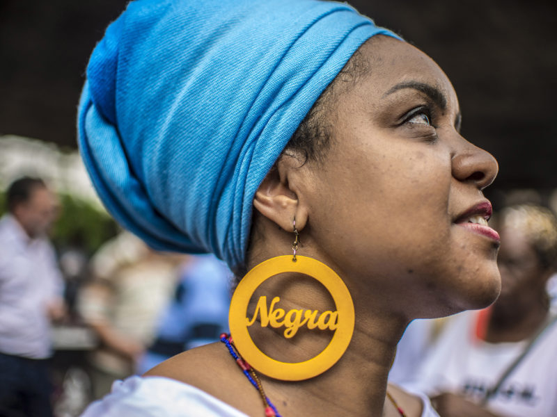 Uma mulher negra de brinco amarelo redondo onde há a palavra "negra" escrita e um turbante azul enrolando todo o cabelo