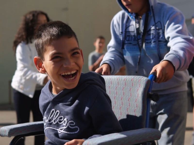 Menino cadeirante está sorridente demais enquanto uma pessoa conduz a cadeira dele
