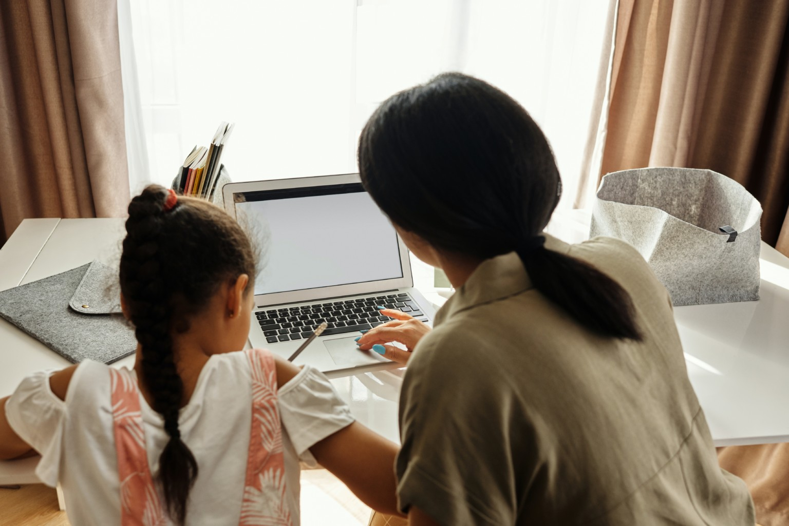 Imagem de uma mulher branca de cabelos escuros, lisos e pretos ensinando algo no notebook para uma menina que está ao seu lado com um lápis na mão. As duas estão de costas para quem bateu a foto