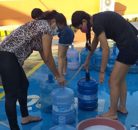 Duas mulheres estão repartindo água, passando um pouco de um galão para outro através de uma mangueira