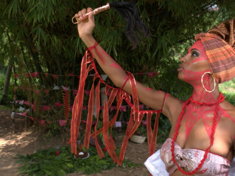 Uma mulher negra está de top tomara que caia, brincos enormes de argola, turbante envolvendo os cabelos, um colar vermelho, tintura vermelha espalhada pelo rosto e lantejoulas também vermelhas penduradas pelo braço direito. Ela está olhando seriamente para um objeto que segura com a mão direita, com o braço levantado acima da altura de sua cabeça