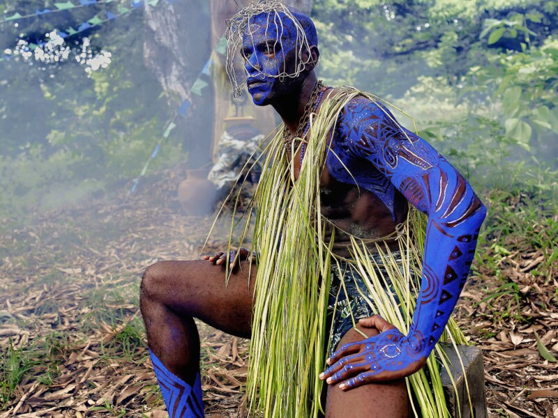 Homem negro de musculatura bem avantajada está ajoelhado em meio a um matagal e com a parte superior do corpo coberta por tinta azul, incluindo o rosto. Ele também está olhando fixamente para algo que não conseguimos saber o que é e está coberto por mato verde sobre a cabeça e o tronco