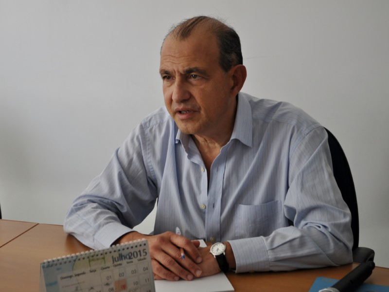 Um homem branco, calvo, de camisa social de mangas compridas e na cor azul, está sentado com as duas mãos sobre a mesa à sua frente