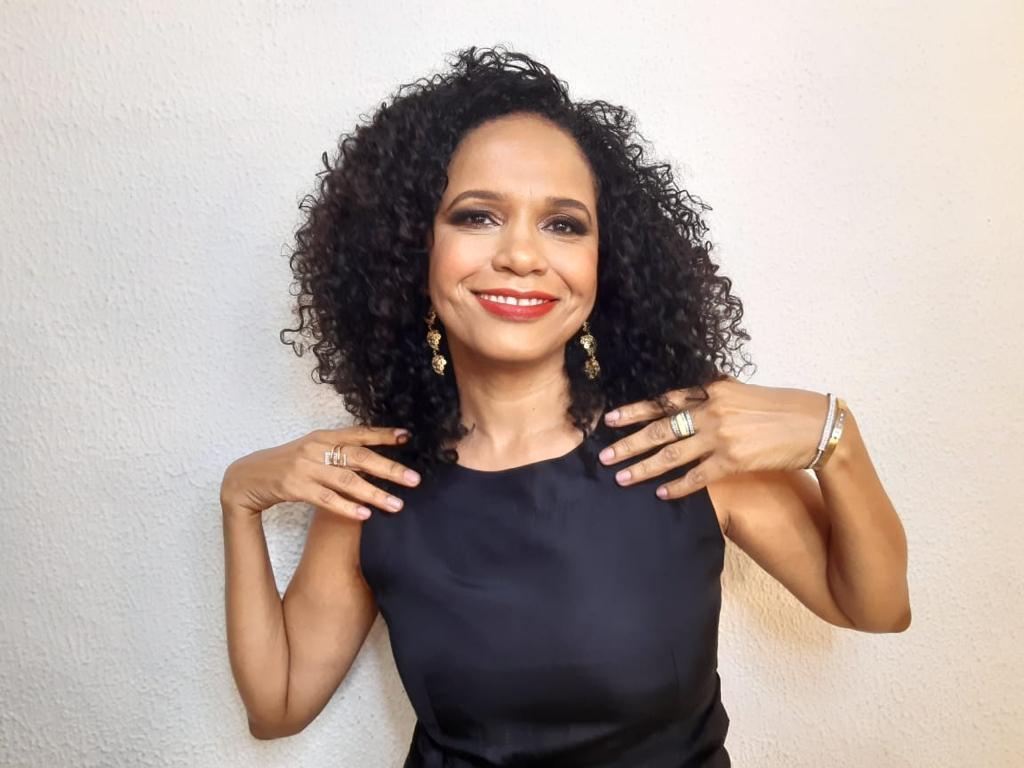 Mulher negra de cabelos cacheados, batom vermelho e brincos chiques está sorridente e trajando um vestido preto