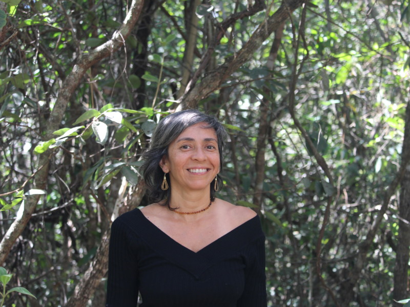 Imagem de uma mulher à frente de várias árvores. Ela está sorrindo e olhando para quem bateu a foto e tem a pele morena, cabelos acinzentados