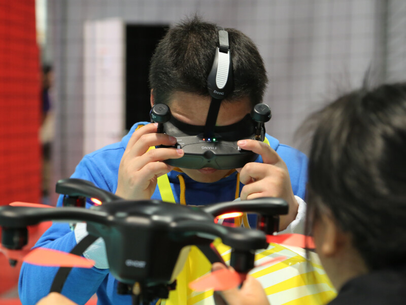Um homem está usando o que parece ser um óculos de realidade virtual e olhando pra baixo. à frente dele e de costas para quem bateu a foto há uma mulher segurando um drone