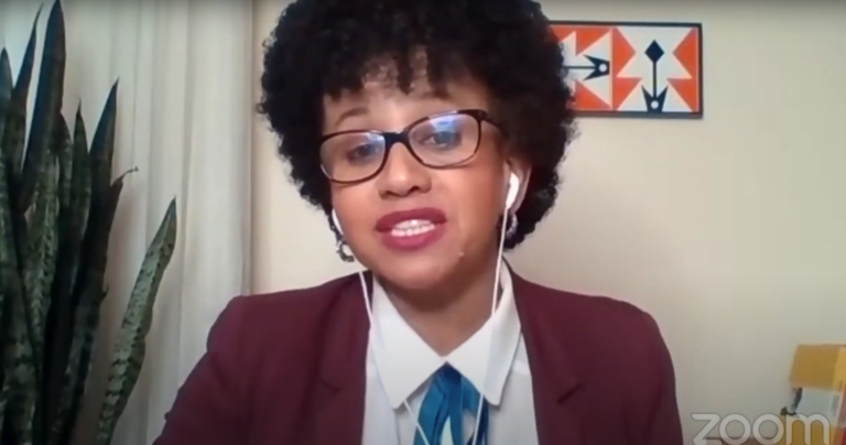 Print de uma tela mostrando uma mulher negra de cabelo black power, óculos de grau e paletó da cor vinho