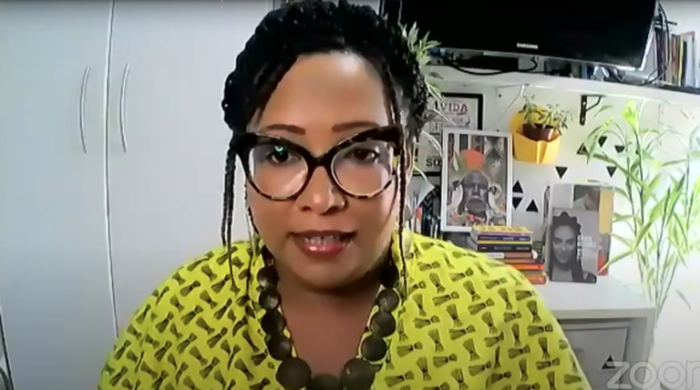 Print de uma tela mostrando uma mulher negra de óculos de grau, colar grosso e blusa amarela